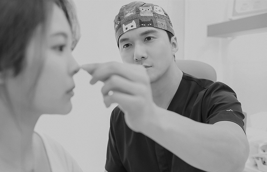 수술모를 쓴 의사가 환자의 코를 짚어 보는 상담 장면(흑백)